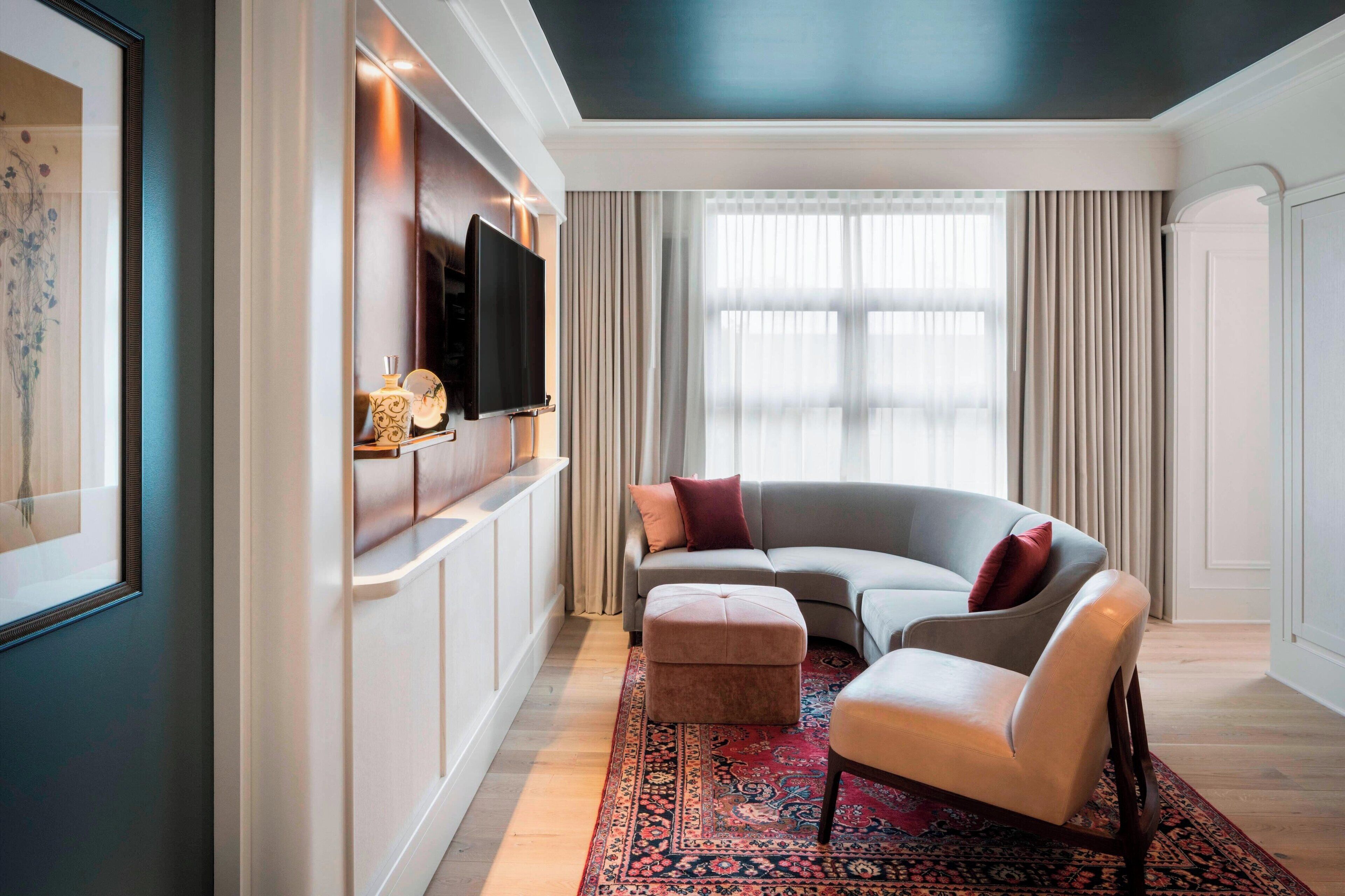 A modern living room with a curved grey sofa, a white armchair, and a red patterned rug. A flat-screen TV is mounted on the wall, and there is a window with sheer curtains.