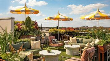 Un patio exterior con varias zonas de estar, incluyendo sofás y sillones, rodeado de plantas en macetas y sombrillas.