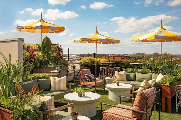 An outdoor patio with multiple seating areas, including sofas and chairs, surrounded by potted plants and umbrellas.