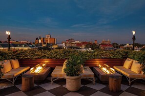 A rooftop terrace with outdoor seating and a fire feature, overlooking a cityscape at dusk.