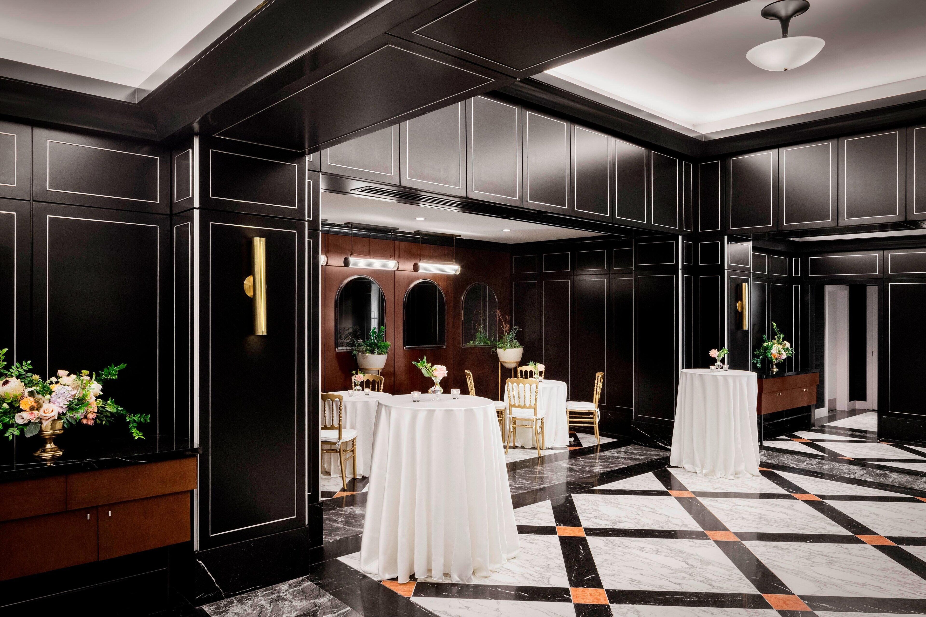 A spacious hotel lobby with a checkered marble floor, black paneling, and round tables with white tablecloths.