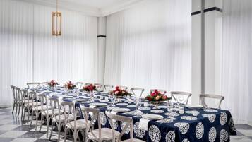 Un espace repas avec des tables rondes disposées pour accueillir six personnes, chacune étant recouverte d’une nappe en dentelle blanche et agrémentée d’un arrangement floral.