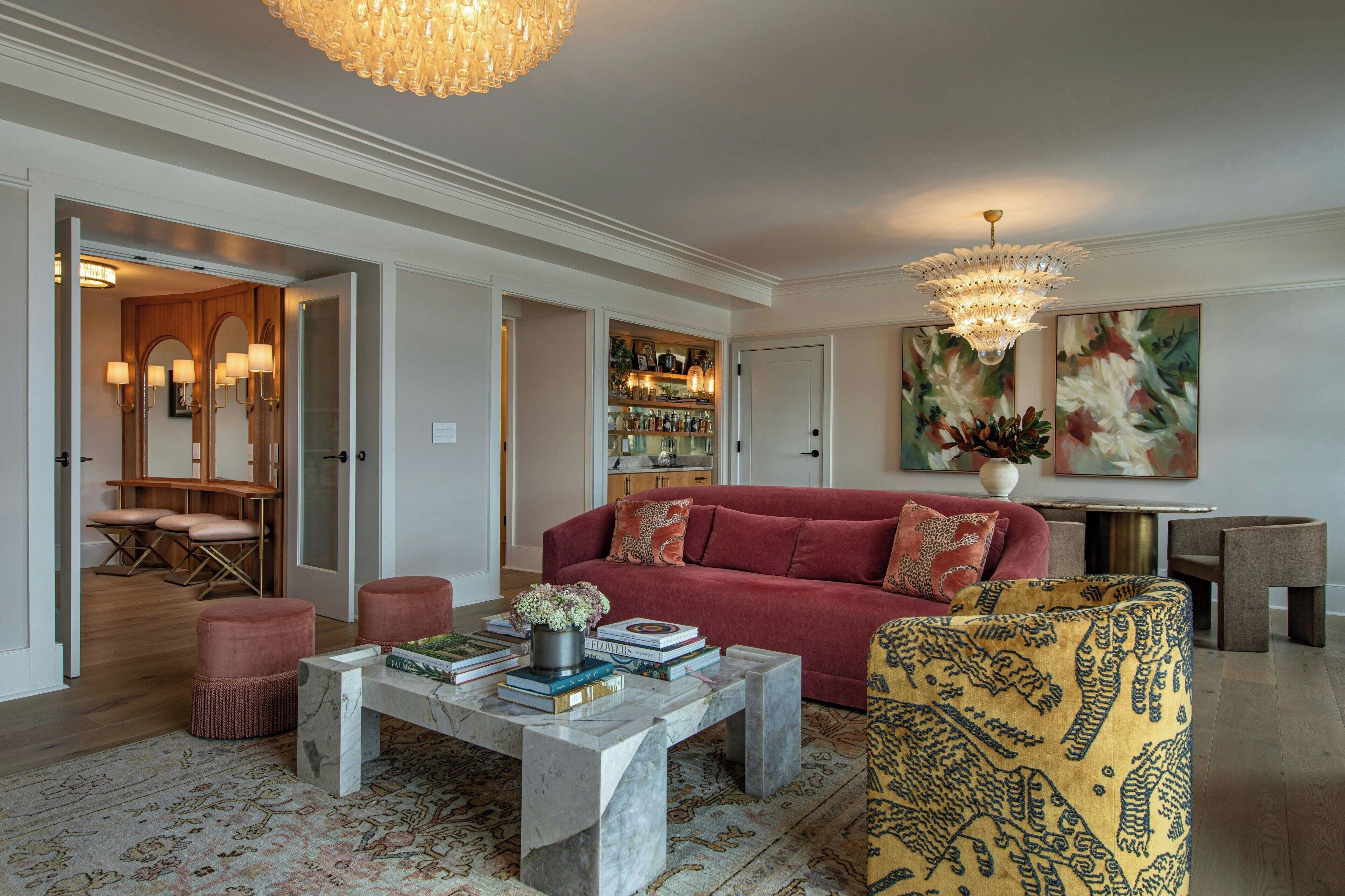 Ein Wohnzimmer mit rotem Sofa, einem Marmortisch und einem gemusterten Sessel. Über dem Sofa hängt eine Chandelieber und an der Wand ein Gemälde.