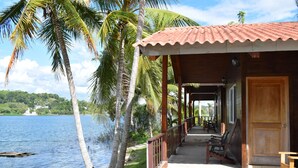 A wooden building with a tiled roof, a porch with benches, and a view of a lake with palm trees. - Hotel Santa Bárbara Tikal (San Benito)