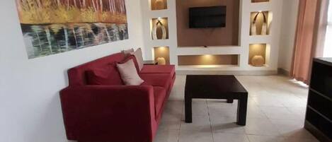 Una sala de estar moderna con un sofá rojo, una mesa de centro, un televisor de pantalla plana montado en la pared, iluminación empotrada y un cuadro grande en la pared.