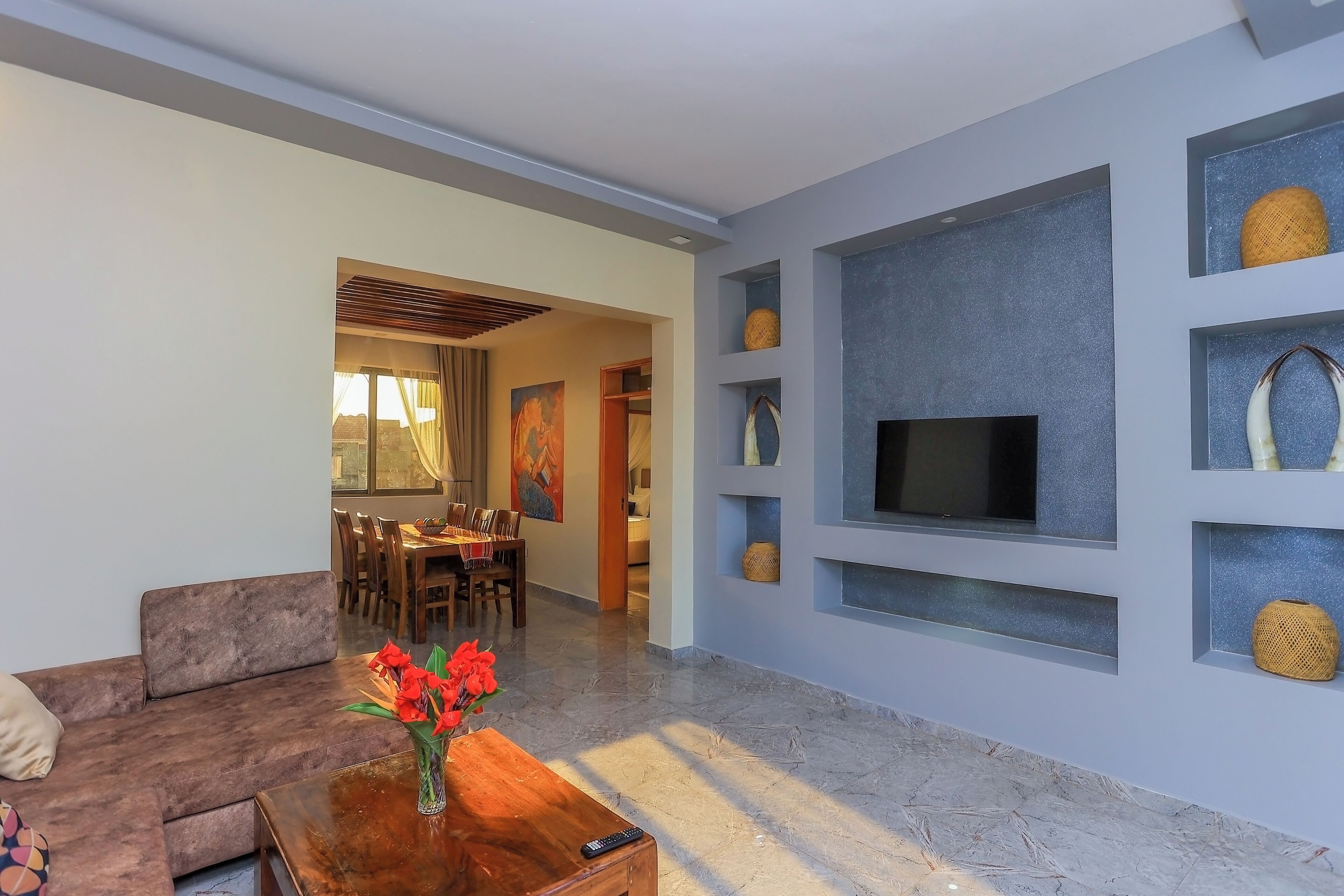 A modern living room with a sofa, a coffee table with a vase, a TV mounted on a wall, and a dining area in the background.
