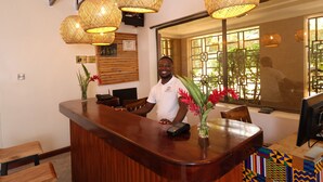 A reception desk with a wooden counter, a digital payment terminal, and a vase with flowers. There are wicker lamps above the desk and a window with a view of greenery outside. - Bon Bini Residence (Kampala)