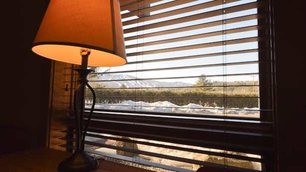 A lamp on a wooden table next to a window with horizontal blinds, offering a view of a snowy landscape with a mountain in the distance. - Timber Inn Motel (Ludlow)