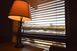 A lamp on a wooden table next to a window with horizontal blinds, offering a view of a snowy landscape with a mountain in the distance. - Timber Inn Motel (Ludlow)