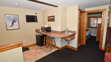 A compact kitchen with a microwave, sink, and stools. A television is mounted on the wall, and there are framed pictures.