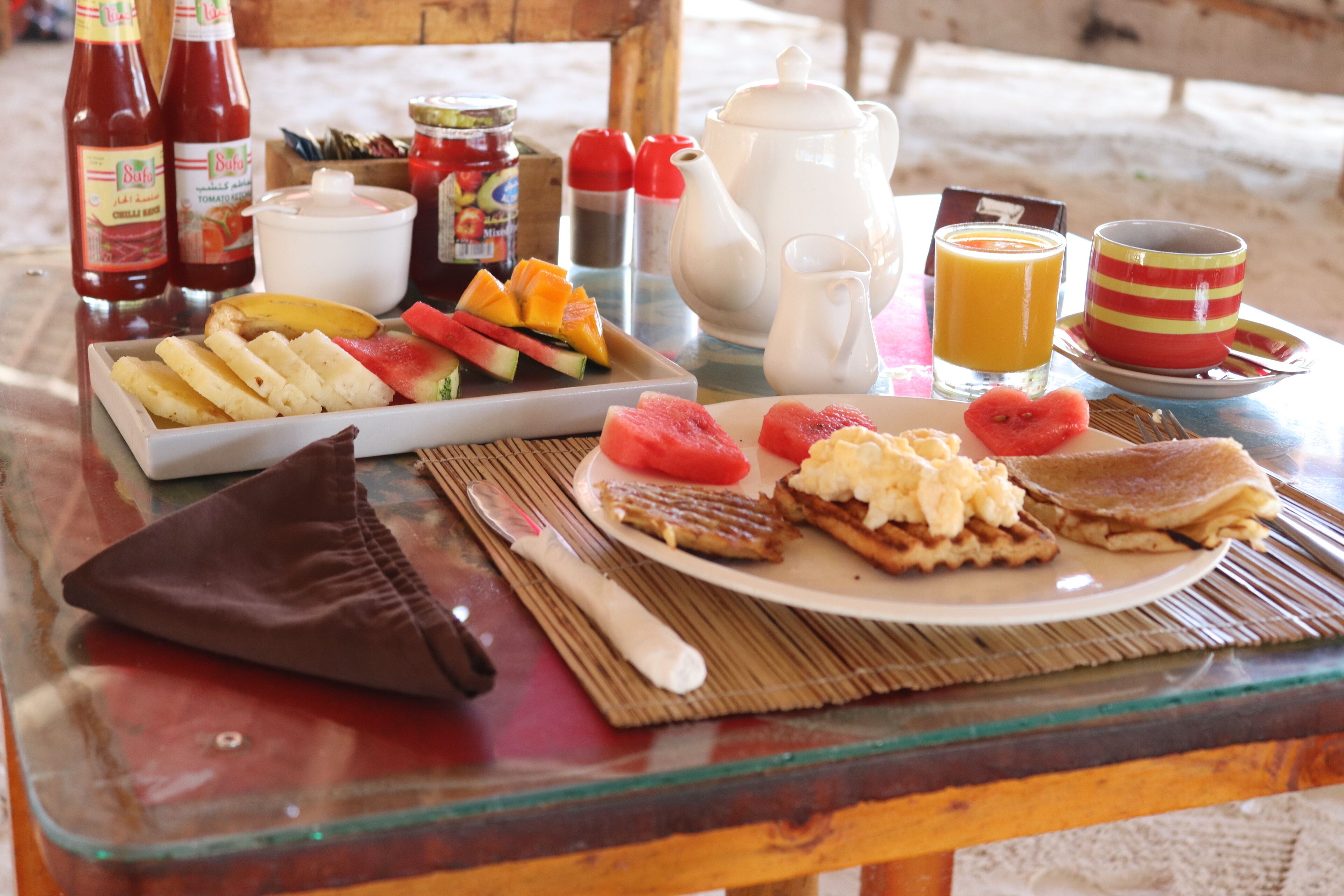 A breakfast table with various food items, a bottle of ketchup, a teapot, a jug of juice, and a cup.
