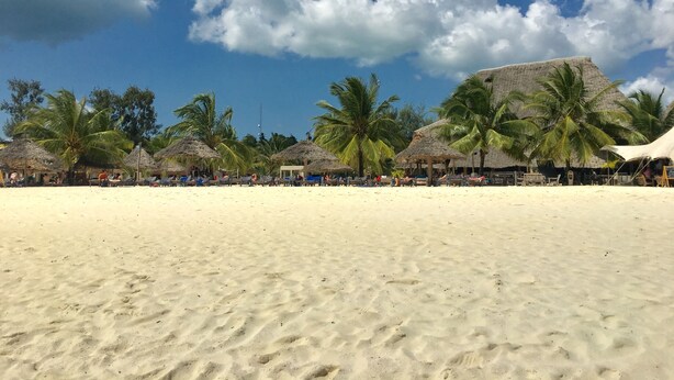 A beach with thatched-roof huts, palm trees, and a large building in the background. - Mocco Beach Villa (Kendwa)