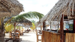 A beachside bar with thatched roofs, wooden tables, and chairs, set against a sandy backdrop with palm trees. - Mocco Beach Villa (Kendwa)