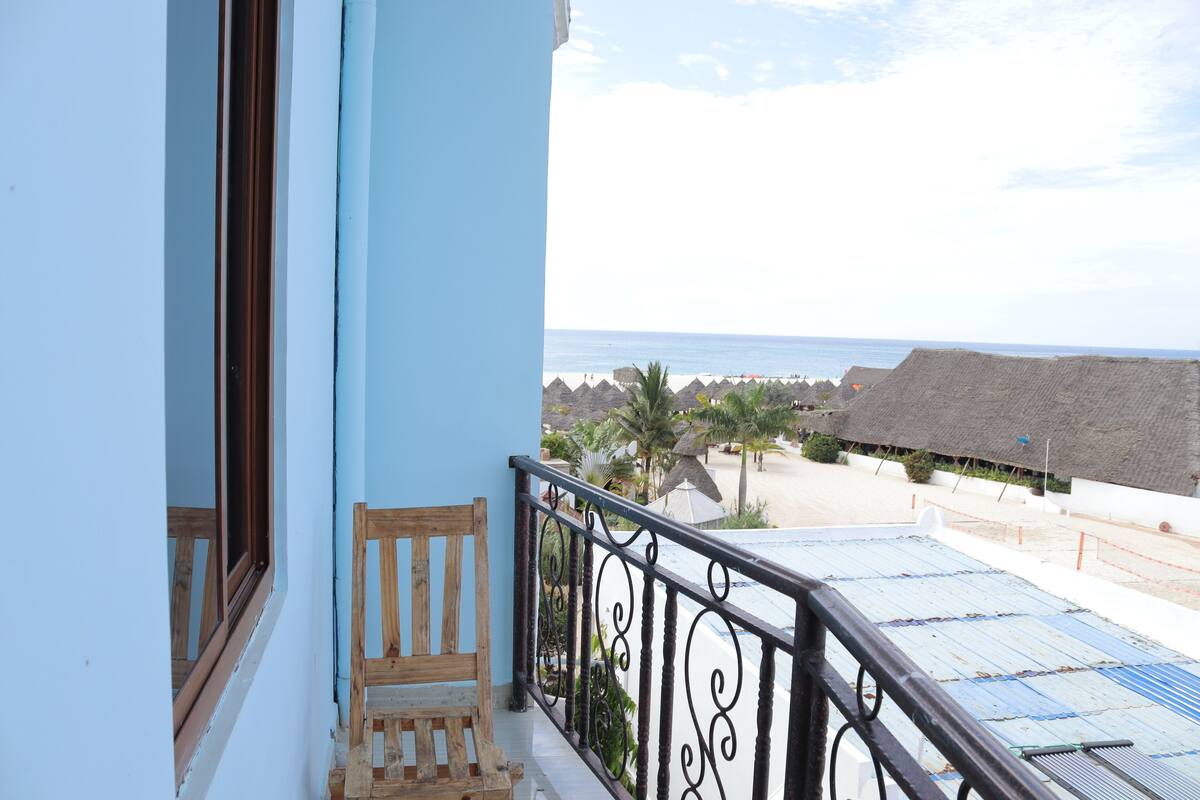 Un balcón con banco de madera, barandilla de metal y vista a la playa con construcciones de techo de paja.