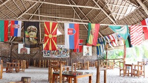 A covered dining area with multiple wooden tables and chairs, flags hanging from the ceiling, and a thatched roof structure. - Mocco Beach Villa (Kendwa)