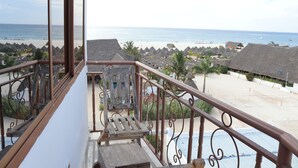 A balcony with a view of a beach, palm trees, and thatched-roof buildings. - Mocco Beach Villa (Kendwa)