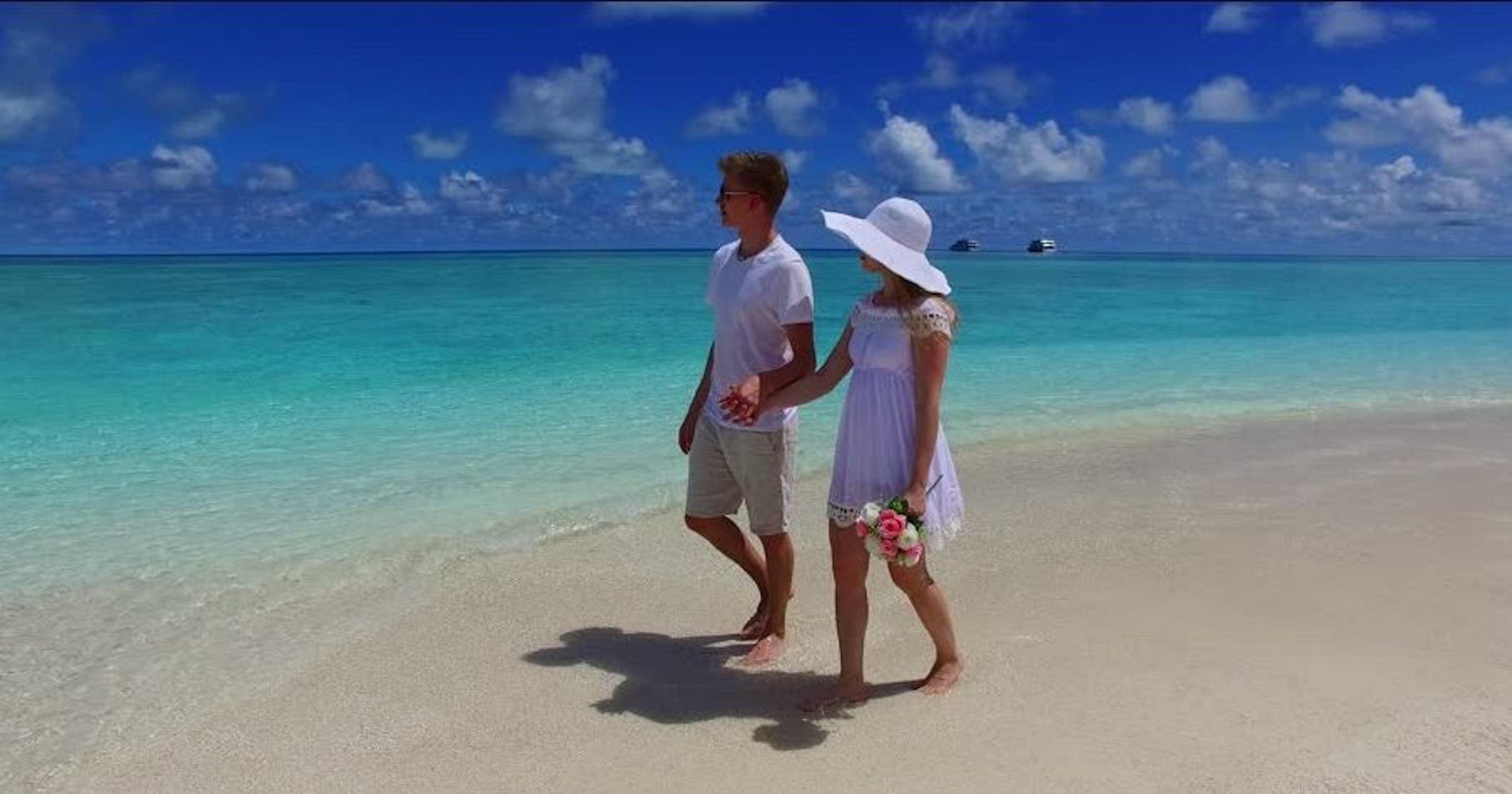 Ein Paar steht an einem Strand mit klarem türkisblauem Wasser und ein paar Booten in der Ferne.