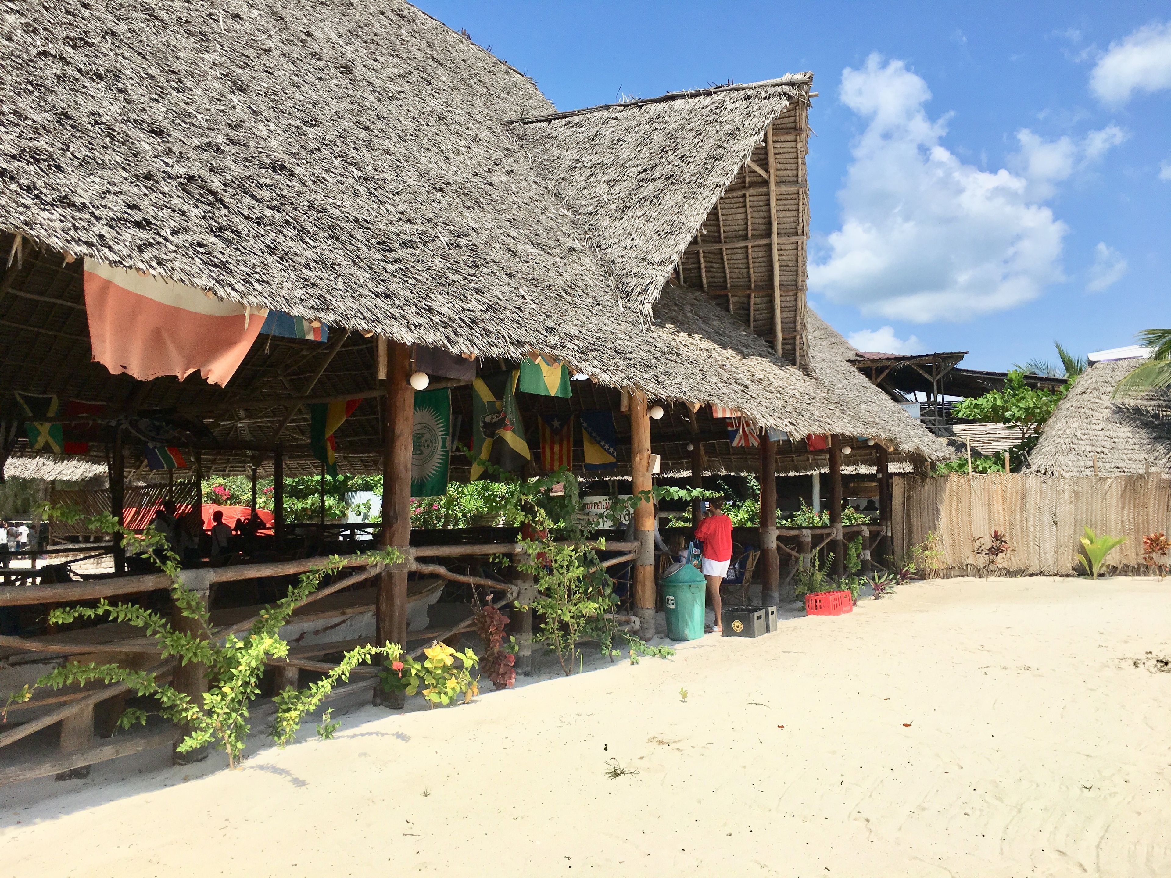 Ein Gebäude am Strand mit Reetdächern und Holzbalken, umgeben von Grünpflanzen und Flaggen.