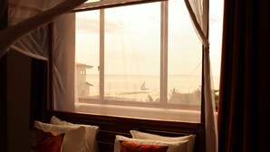 A room with a bed, a window with curtains, and a view of the sea and buildings. - Mocco Beach Villa (Kendwa)
