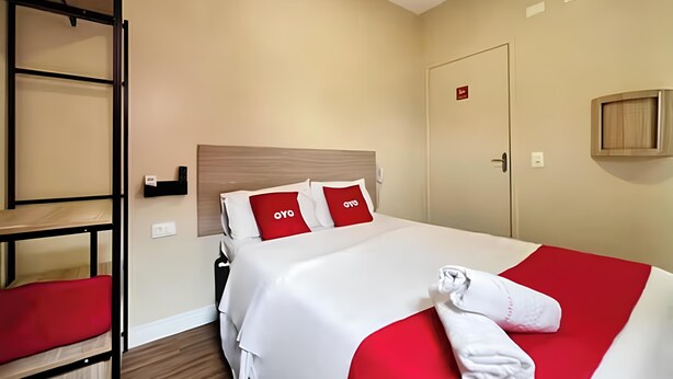 A hotel room with a bunk bed, a large bed with red and white bedding, a towel on the bed, and a door with a sign. - OYO Hotel Itarantim, São Paulo (São Paulo)