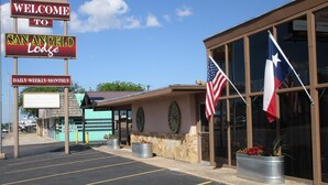Entrada de hotel con un letrero que dice 'Welcome to San Angelo Lodge', una bandera estadounidense, una bandera de Texas y un estacionamiento.