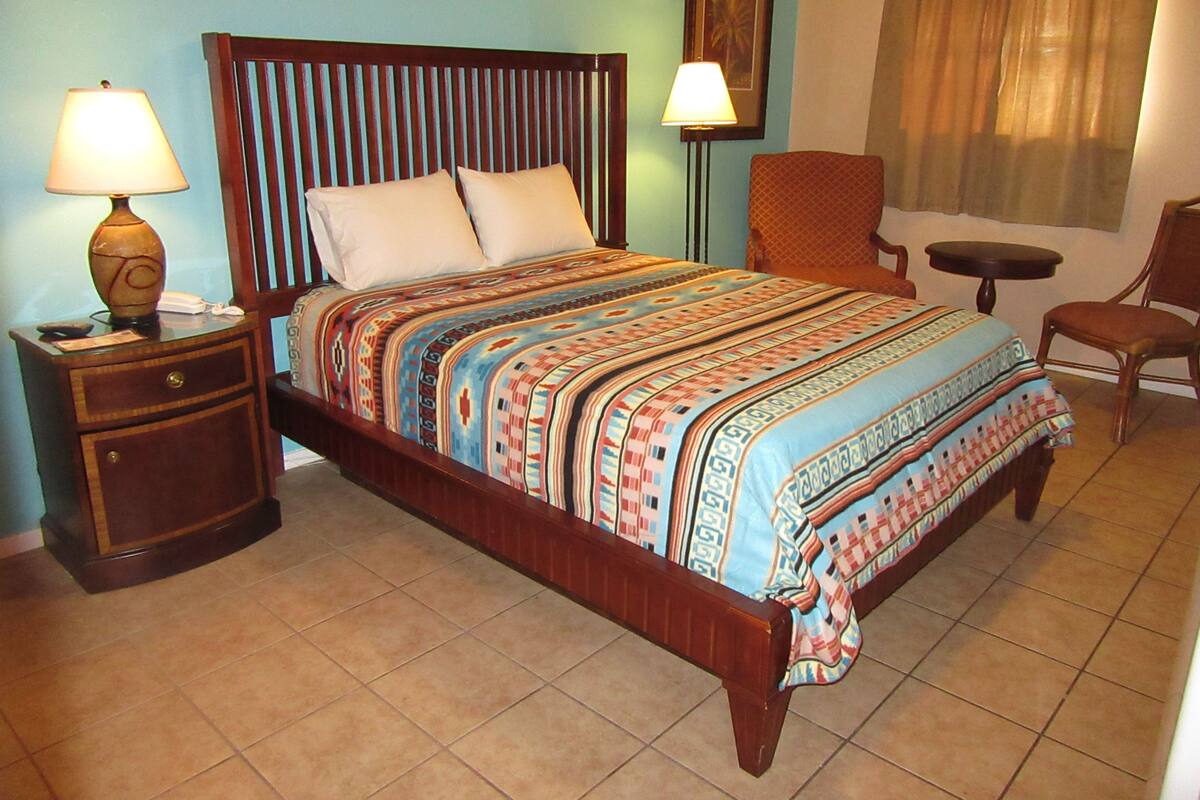 A hotel room with a wooden bed, a nightstand, a chair, a small table, and a lamp.