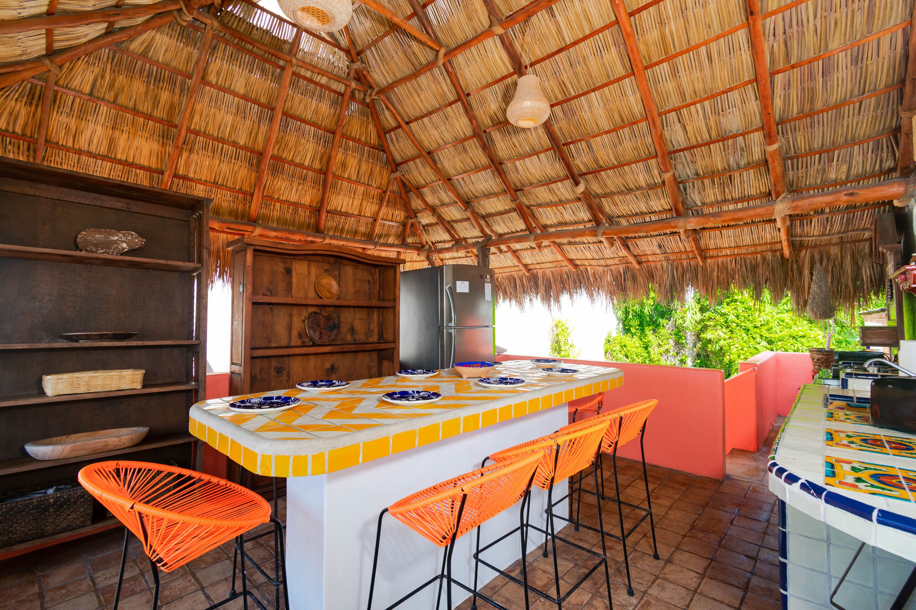 Zona de bar con techo de paja, una barra central, taburetes naranjas y un revoque de azulejos.