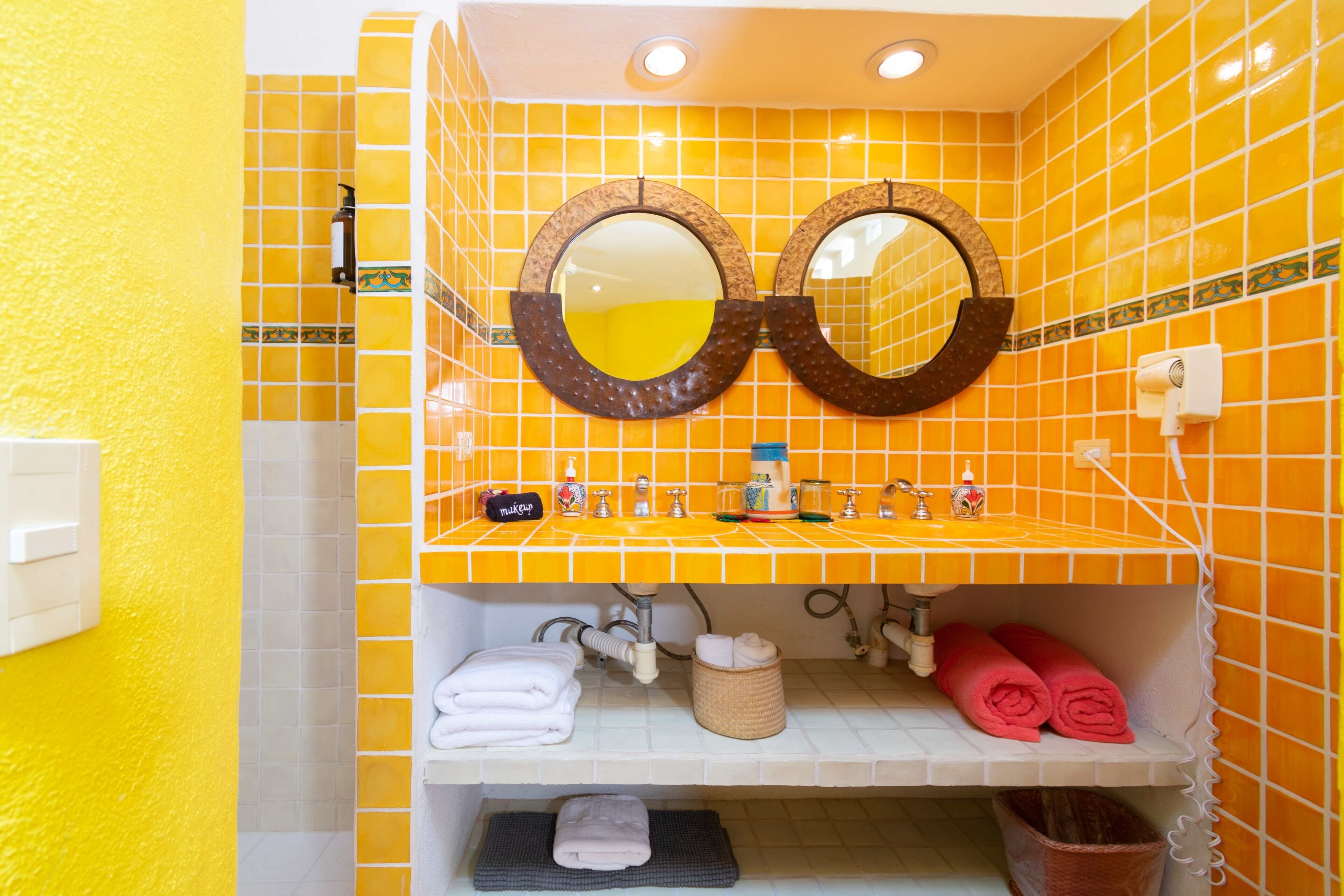 A bathroom with yellow tiles, two round mirrors, a sink with toiletries, and towels on shelves.