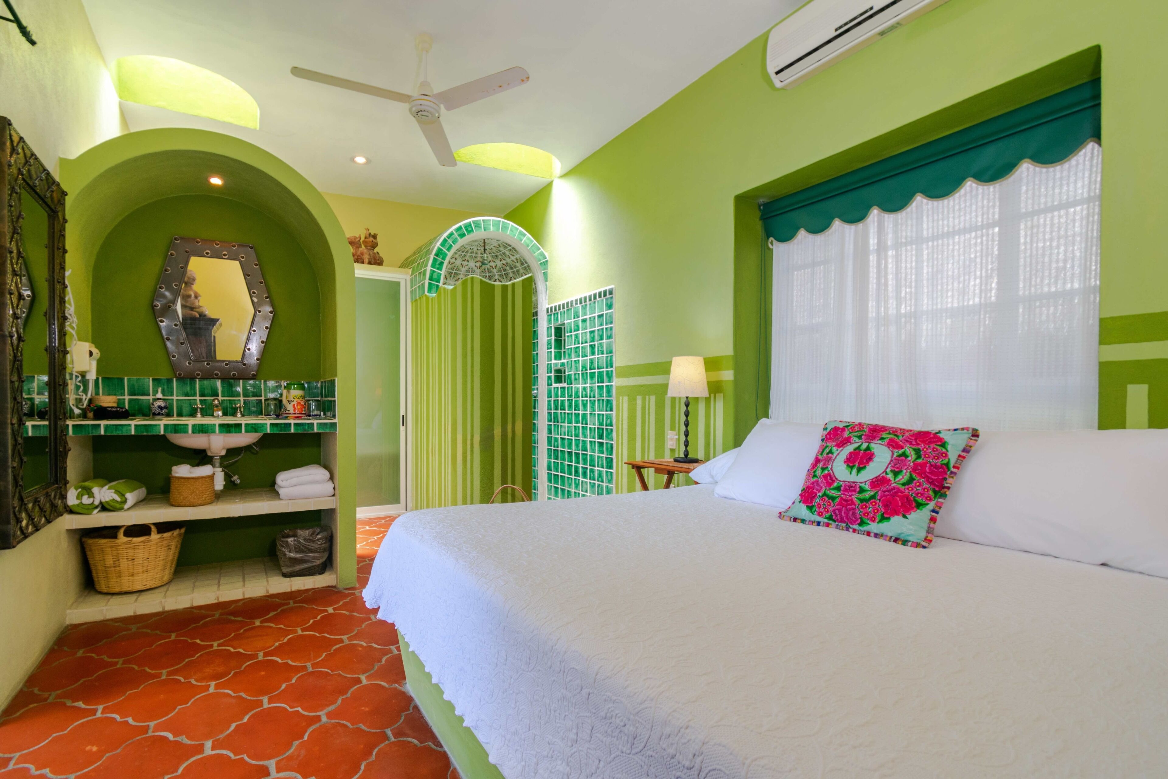 Un dormitorio con cama, mesita de noche, lámpara, espejo, pared verde, ventilador de techo y ventana con cortina.