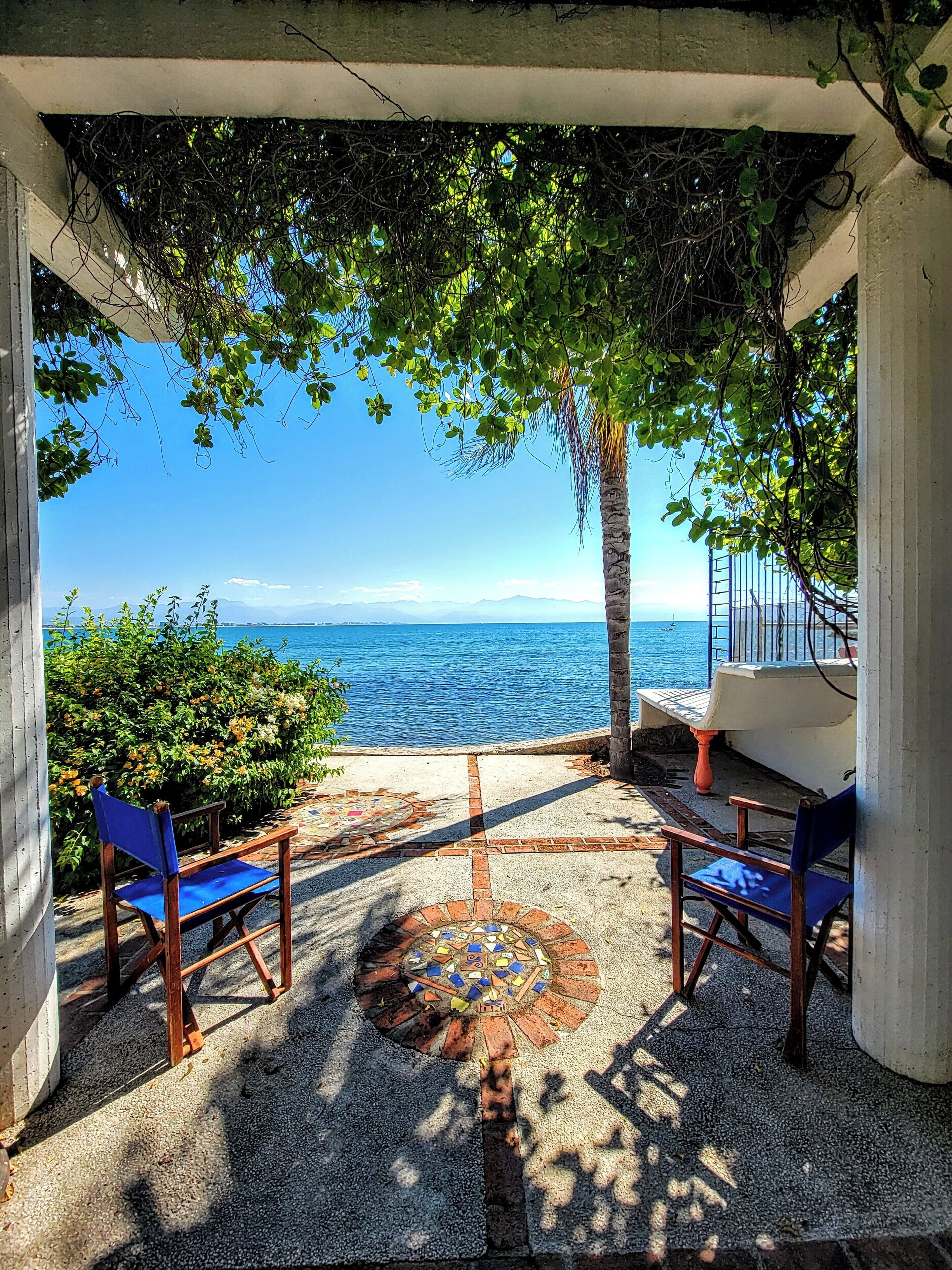 Uma área de estar ao ar livre com vista para o mar, com piso de azulejos e cadeiras de madeira.