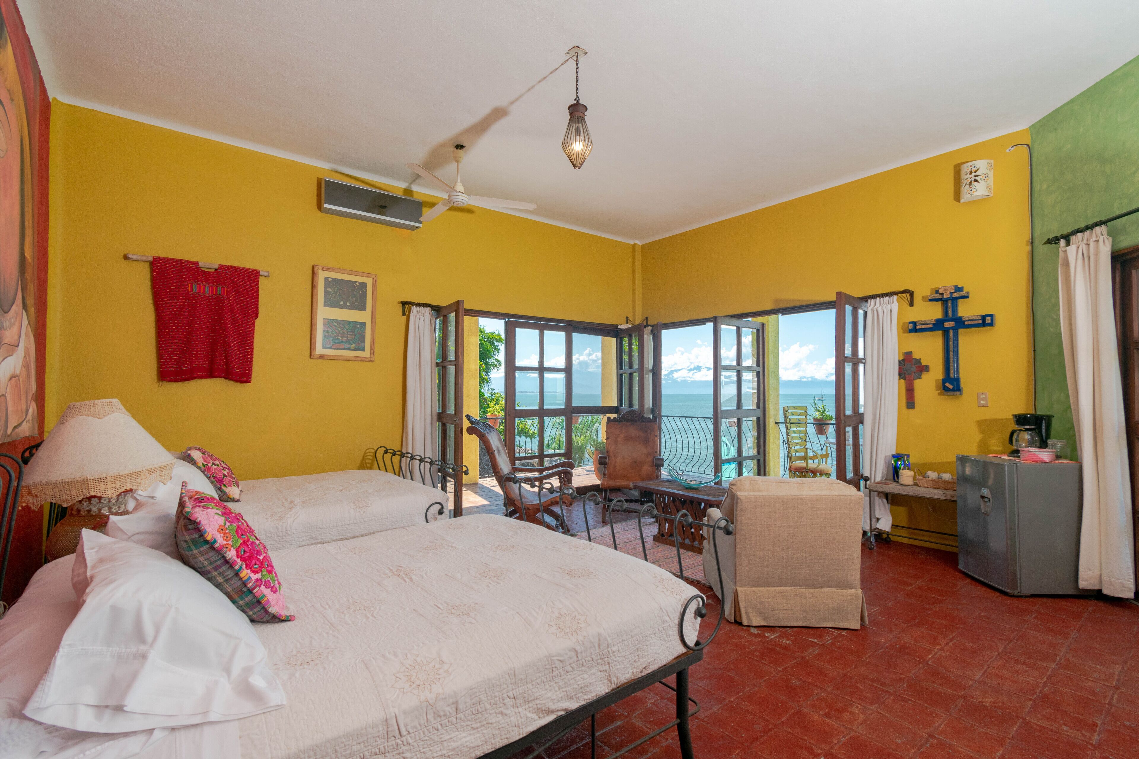 A room with two beds, a ceiling fan, a view of the sea, a small table with chairs, and a refrigerator.
