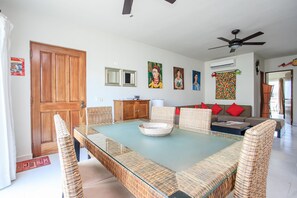 A dining area with a glass-top table, wicker chairs, a wooden cabinet, and a sofa with red cushions.