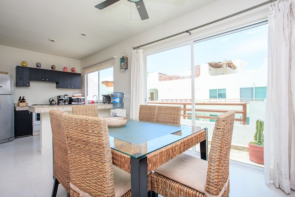 A modern kitchen with dark cabinets, a glass-top dining table, wicker chairs, and a large sliding glass door leading to a balcony with a view of buildings and a cactus plant.