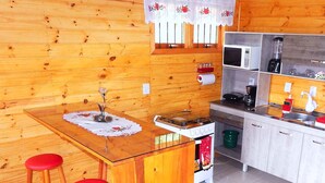 Interior de madeira com cozinha, incluindo fogão, micro-ondas e pia. Duas cadeiras vermelhas em uma mesa de madeira com toalha de renda.