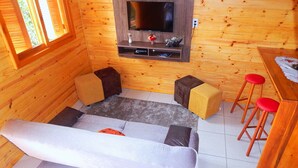 Interior acolhedor de uma cabana com um sofá, dois banquinhos, uma TV fixada em uma parede de madeira e uma mesa com um banquinho vermelho.
