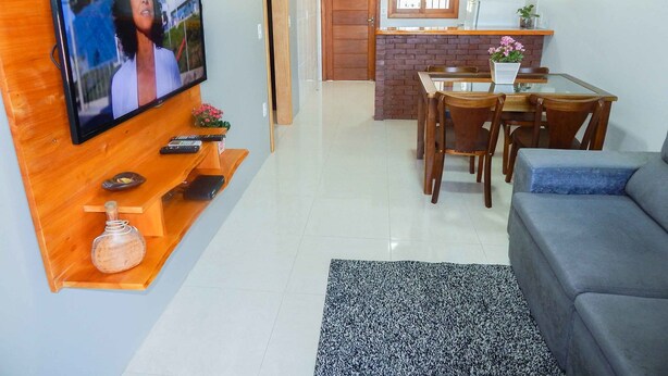 A modern living room with a flat-screen TV, wooden furniture, a dining area with a brick accent wall, and a kitchen with wooden cabinets. - Residencial de Luxe Ouro 01 (Canela)