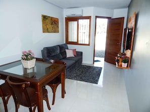 A living room with a sofa, a glass dining table, wooden chairs, a small potted plant, a painting, and a door leading outside. - Residencial de Luxe Ouro 01 (Canela)