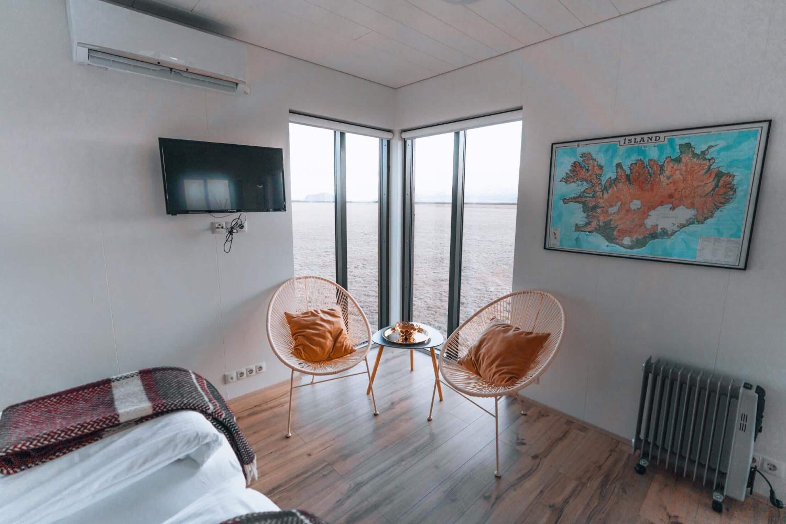 A room with a bed, two wicker chairs, a small table, a television, a radiator, and a large window with a view of the sea.