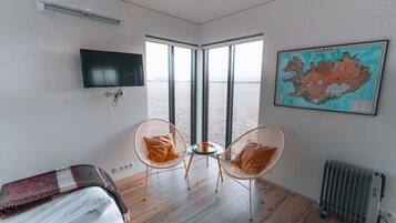 Une chambre avec un lit, deux fauteuils en osier, une petite table, un téléviseur, un radiateur et une grande fenêtre donnant sur la mer.