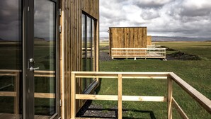 Un edificio de madera con puerta de cristal y una terraza de madera que conduce a una estructura de madera en un campo cubierto de hierba.