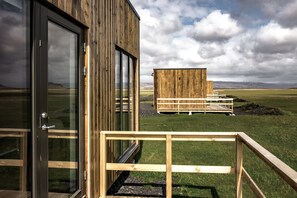 A wooden building with a glass door and a wooden deck leading to a wooden structure in a grassy field. - Brú Guesthouse (Rangárþing eystra)