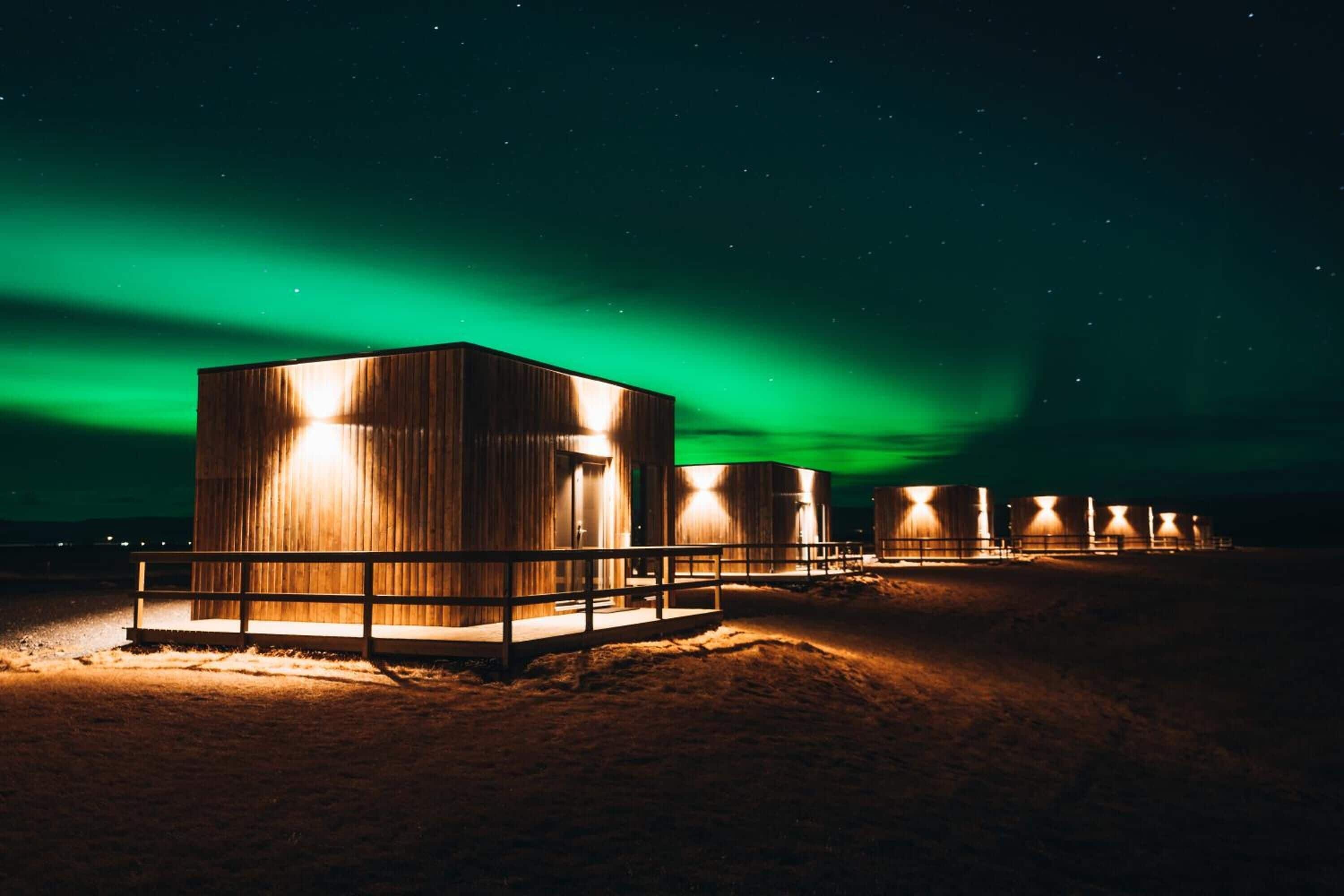 A row of wooden cabins with a metal railing in front, set against a night sky with green aurora.