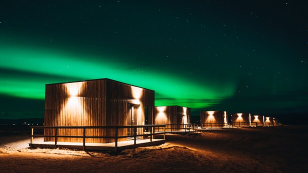 Filas de cabañas de madera con un porche de madera al frente, bajo un cielo nocturno con aurora boreal verde.