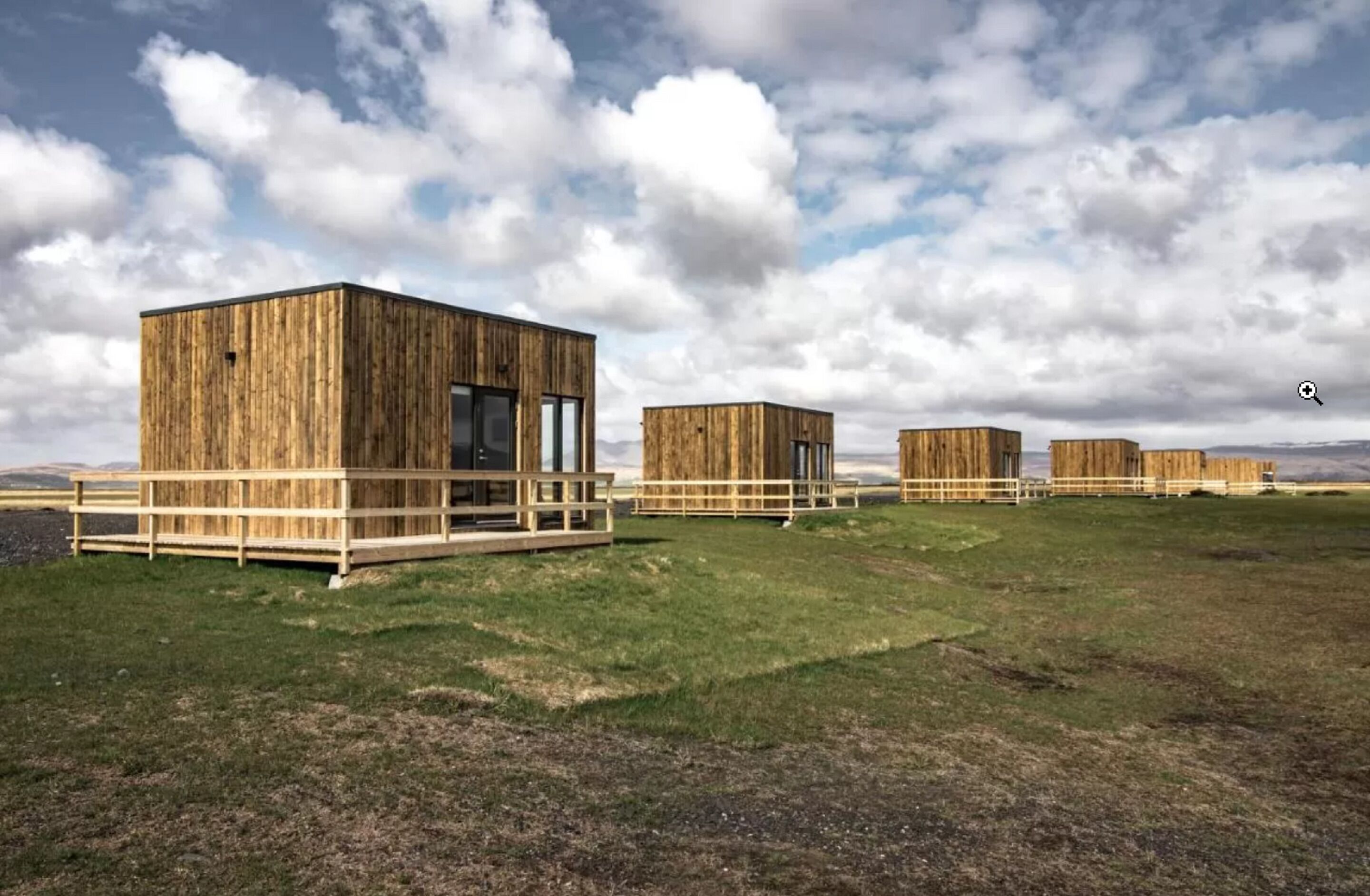 A row of wooden cabins with a deck, set against a grassy field and a cloudy sky.