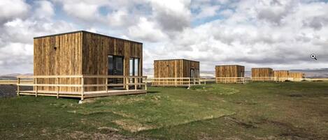 A row of wooden cabins with a deck, set against a grassy field and a cloudy sky.