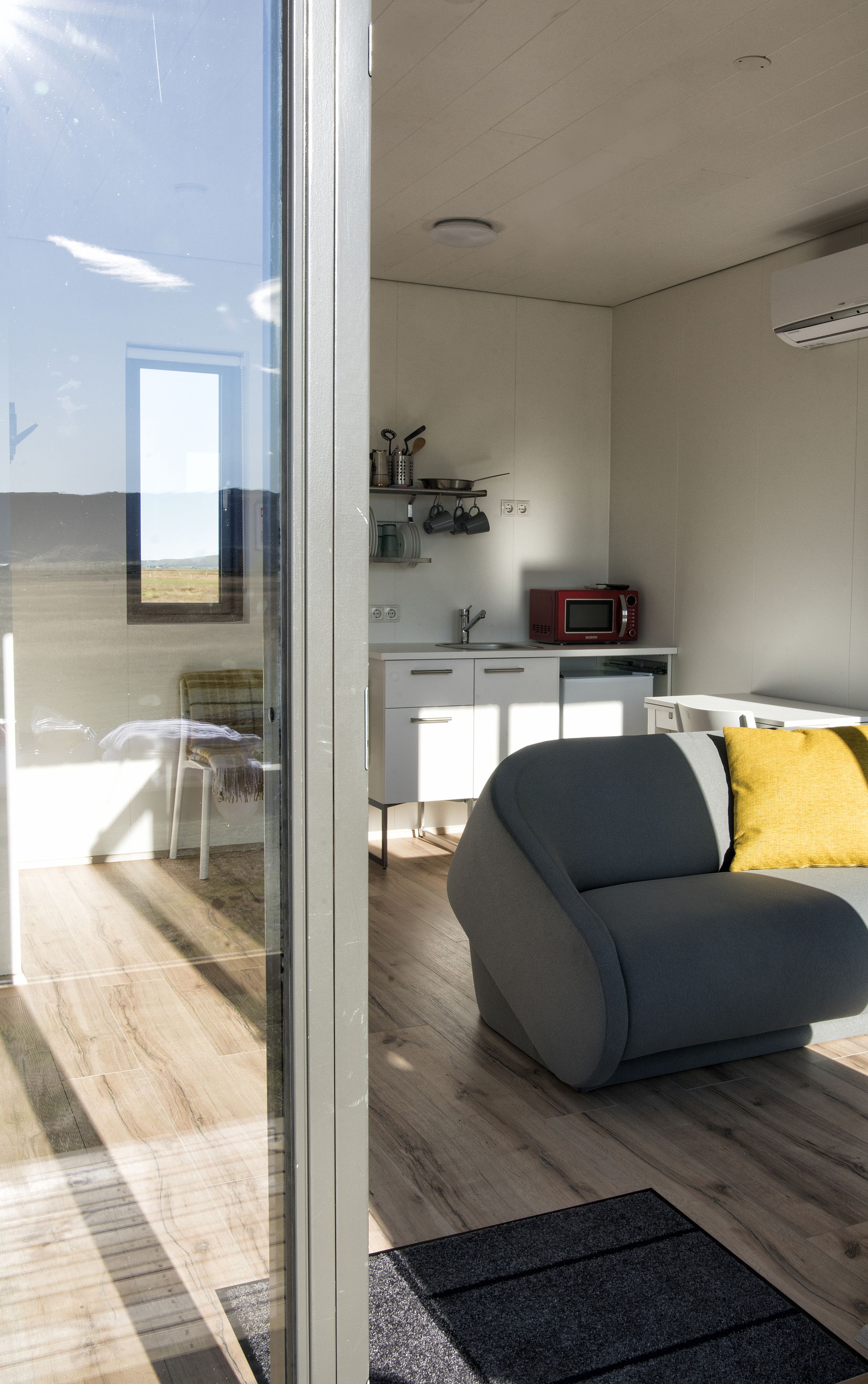 Une cuisine moderne avec des meubles blancs, un four à micro-ondes et un évier. Le salon est doté d’un canapé gris agrémenté d’un coussin jaune.