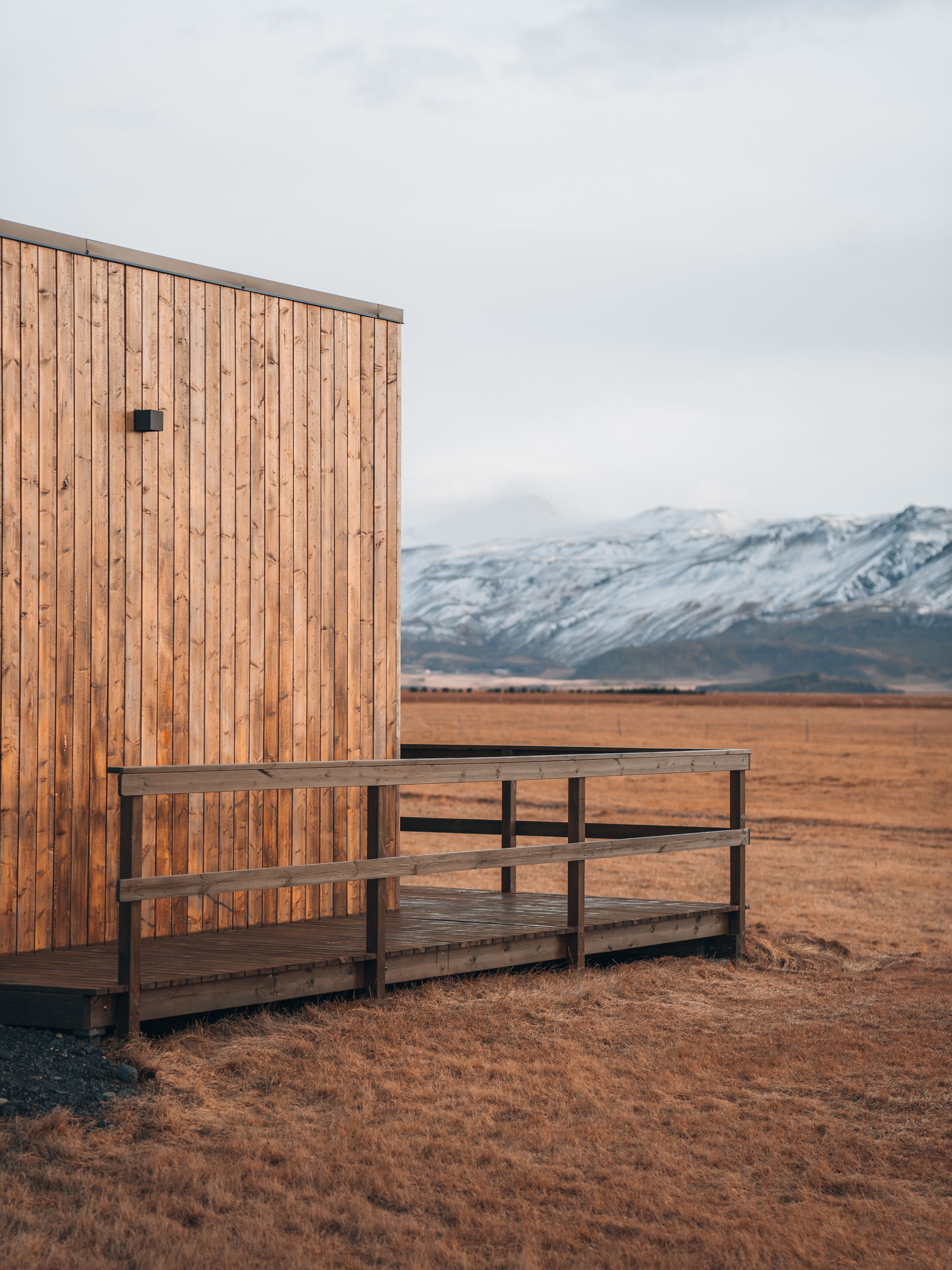 A wooden cabin with a bench in front, set against a backdrop of snow-capped mountains and a cloudy sky.