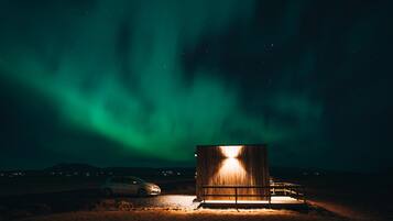 A wooden cabin with a bench and a car parked outside, under a night sky with visible stars and aurora borealis.