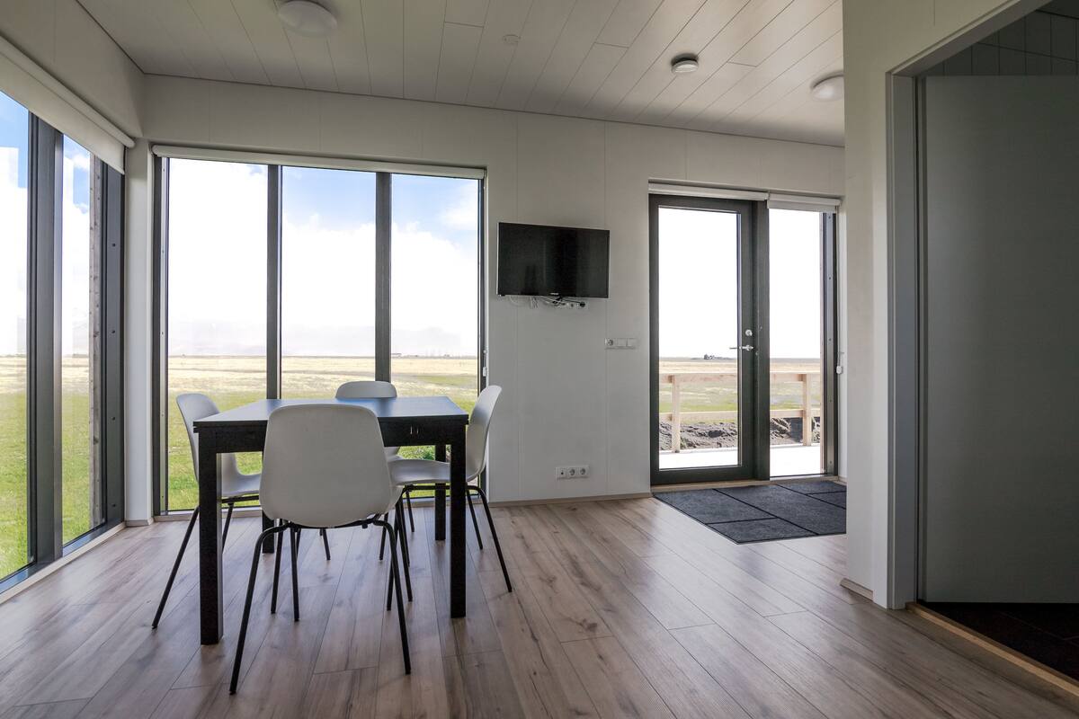 A modern dining area with a table and chairs, large windows, and a flat-screen TV mounted on the wall.
