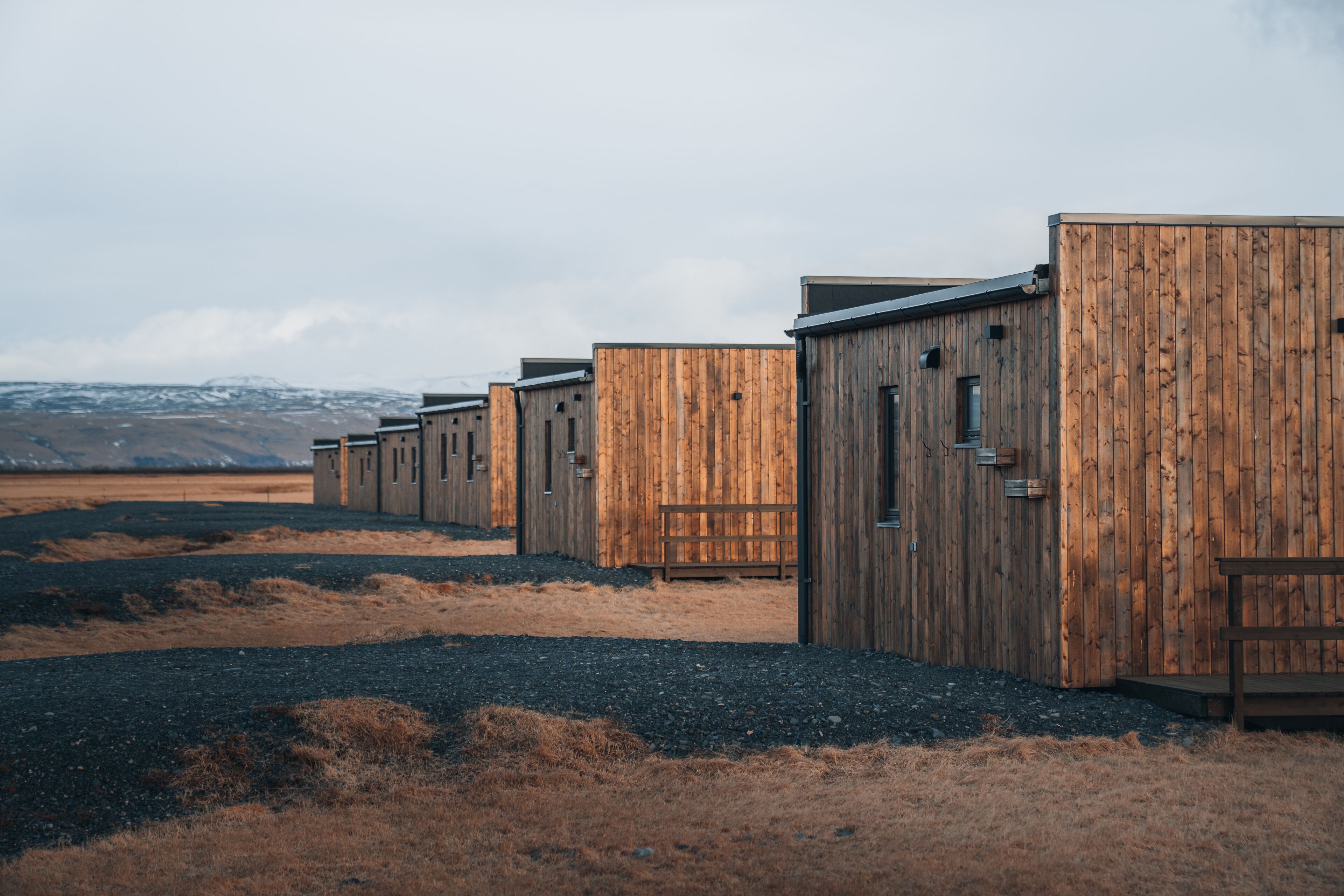 A row of wooden cabins with a gravel path leading to them, set against a mountainous backdrop.